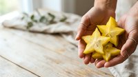 Close-up of hands holding fresh yellow star fruit on wooden surface with herbs in background, suitable for food, cooking, and healthy eating content.