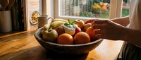 Fresh organic fruits in wooden bowl on kitchen counter with natural sunlight streaming through window creating warm atmosphere for healthy eating lifestyle.
