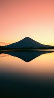 Silhouetted volcanic mountain reflected in still lake water at sunrise with warm peachy sky gradient, serene nature landscape for travel and meditation projects.