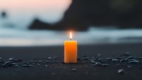 Single lit candle glowing at dusk on rocky beach shore, creating peaceful ambiance against blurred ocean background and wet pebbles.