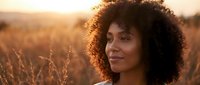 Young African American woman with curly hair smiling peacefully in golden wheat field during sunset, natural beauty and serenity in warm outdoor setting.
