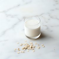Glass of fresh oat milk with scattered raw oats on marble surface, minimalist composition showing dairy alternative beverage in natural light.