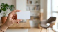 Person holding blank white business card in modern home office with potted plant, wooden desk, and blurred bookshelf background for branding and corporate identity projects.