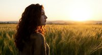 Young woman with curly hair in knit sweater gazing at golden sunset over wheat field during golden hour creating peaceful rural landscape scene.