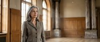 Middle-aged woman with long gray hair and glasses wearing a gray blazer, standing in an elegant empty interior space with wooden doors and arched windows, suitable for corporate, professional, and lifestyle editorial use.
