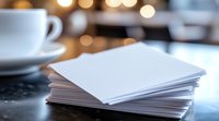 Stack of blank white paper sheets on dark table with coffee cup and warm bokeh lights in background, creating cozy atmosphere for writing or creative work.