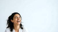 Young woman with dark curly hair and olive complexion wearing white shirt, laughing joyfully against neutral background for portrait and lifestyle use.