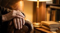 Elderly hands resting on wooden walking cane in warm home interior with books and soft lighting creating peaceful retirement atmosphere.