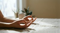 Woman practicing meditation in mudra pose on bed with morning sunlight streaming through window creating peaceful wellness atmosphere for mindfulness and relaxation.