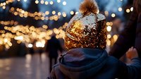 Person wearing gold sequin winter hat with fur pom-pom against blurred holiday lights background, creating festive evening atmosphere.