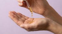 Woman hands applying golden facial oil serum from glass dropper bottle for skincare routine and beauty treatment on pink background.