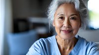 Senior Asian woman with gray hair smiling warmly at camera in modern home interior. Mature female portrait showing confidence and contentment in retirement.
