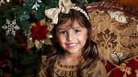 Young Asian girl with bright smile wearing brown dress and bow headband poses near decorated Christmas tree and vintage chair during holiday photo session.