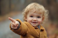 Cheerful toddler with curly blonde hair wearing brown fleece jacket points and smiles outdoors in autumn setting, capturing genuine joy and childhood wonder.