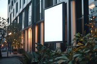 Illuminated blank advertising billboard mockup on modern building exterior with decorative plants and evening city lights, ready for branding display.