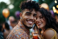 Young Hispanic man in sequined shirt and Latin woman in yellow dress enjoying outdoor party with cocktails and festive bokeh lights and balloons in background.