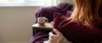 Young woman with long red hair hugging a burgundy pillow at home, cozy living room with coffee cup in background, relaxed mood.