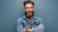 Confident bearded man with glasses and denim jacket smiling with crossed arms on blue background. Professional portrait for business and lifestyle concepts.