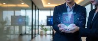 Businessman in dark suit holding digital hologram of circuit board and technology interface in modern office environment with glass partitions.