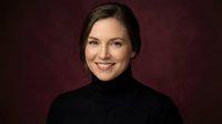 Professional young Caucasian businesswoman in black turtleneck smiling confidently against dark burgundy background for corporate headshot photography.