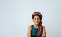 Young woman with South Asian ethnicity wearing a green sleeveless top and pink patterned head wrap, smiling at camera against white background for portrait and lifestyle use.