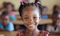 Young African girl with braided hair and pink bows smiling brightly in classroom setting, wearing colorful floral dress. Natural lighting highlights genuine expression.