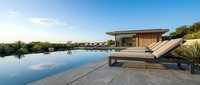 Modern luxury villa with infinity pool and wooden lounge chairs on stone deck. Contemporary architecture with glass walls and natural materials for vacation rental.