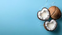 Fresh coconuts cracked open in heart shape with white flesh and splashing milk on vibrant blue background, copy space for summer tropical design.
