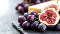 Fresh ripe purple figs cut in half showing juicy red flesh texture alongside dark grapes on rustic stone surface with soft natural lighting and shallow depth of field.