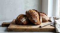 Freshly baked artisan sourdough bread loaves on rustic wooden cutting board by window. Homemade crusty brown bread for bakery, kitchen, cooking concepts.
