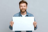 Smiling young man with beard holding blank white poster against gray background, ready for your message or advertisement.