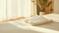 White cozy slippers on bed in morning sunlight with window shadows, minimalist bedroom interior with houseplant and curtains creating peaceful atmosphere.