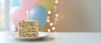 Colorful birthday cake slice with sprinkles and lit candle on white plate against soft pastel balloons and bokeh lights background for celebration.