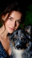 Young Caucasian woman with green eyes and natural makeup embracing Australian shepherd dog with blue merle coat. Close-up portrait against dark background with bokeh effect.