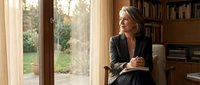 Middle-aged woman with light brown hair, Caucasian ethnicity, wearing dark blazer, sitting in armchair with notebook in home library, looking toward window with garden view.