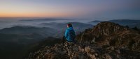 Hiker in blue jacket standing on rocky mountain peak at sunrise overlooking misty valleys and distant ridges. Adventure and outdoor exploration.