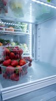 Fresh strawberries in plastic containers stored inside open refrigerator with vegetables on shelves. Cold storage preserves fruit freshness and quality.