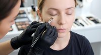 Professional eyebrow artist applying permanent makeup with precision tool on young Caucasian woman client in modern beauty salon studio.