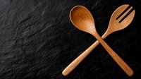Crossed wooden spoon and fork on dark stone background. Traditional handcrafted kitchen utensils made from natural bamboo material for cooking and serving.