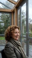 Middle-aged woman with reddish-brown curly hair and fair complexion, wearing a brown blazer and gray scarf, smiling while seated indoors near large modern windows overlooking garden.