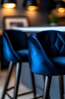 Modern blue velvet bar stools with quilted pattern against blurred restaurant background, warm ambient lighting creates luxurious atmosphere for hospitality settings.