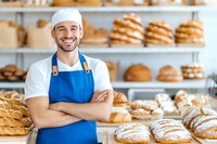 Confident male baker in blue apron standing with crossed arms in artisanal bakery, surrounded by fresh bread loaves and pastries on wooden shelves.