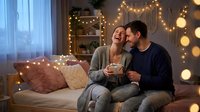 Happy couple enjoying hot drinks together on cozy sofa with warm string lights creating romantic atmosphere at home during evening relaxation time.