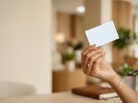 Cropped hand of woman holding blank white card in modern home interior with blurred background, suitable for business card mockups, promotional campaigns, and marketing materials.