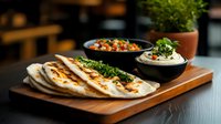 Fresh grilled naan bread served on wooden board with hummus and spicy chickpea curry dips, garnished with fresh herbs and microgreens for restaurant menu.