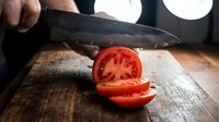Caucasian male chef's hands slicing fresh red tomato with sharp knife on wooden cutting board in professional kitchen with studio lighting.