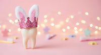 White tooth with bunny ears and pink glitter crown on soft pink background with warm bokeh lights for dental care and Easter celebration concepts.