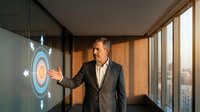 Middle-aged Caucasian businessman in suit presenting holographic target interface in modern office with large windows during golden hour lighting.