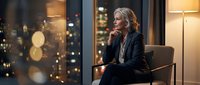 Mature woman with blonde hair and fair complexion, wearing professional dark blazer, sitting thoughtfully by window with city lights bokeh in background at dusk, suitable for corporate, business leadership, and professional development concepts.