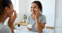 Young mixed-race woman applying face cream in bathroom mirror, smiling while doing morning skincare routine with moisturizer jar on counter.